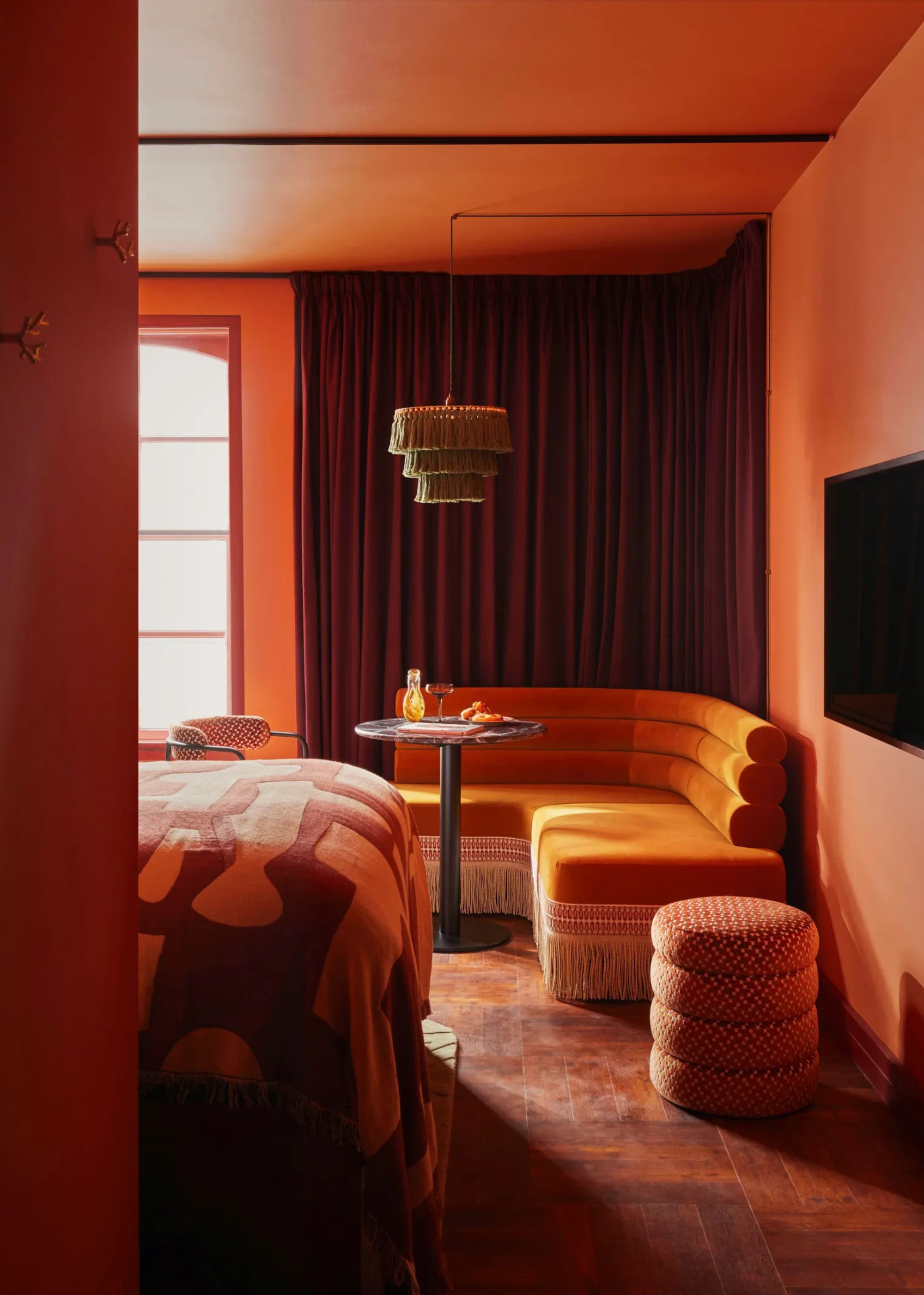 A 70s revival style bedroom with banquette fringe seating, patterned ottomans, a TV, and a fringe pendant