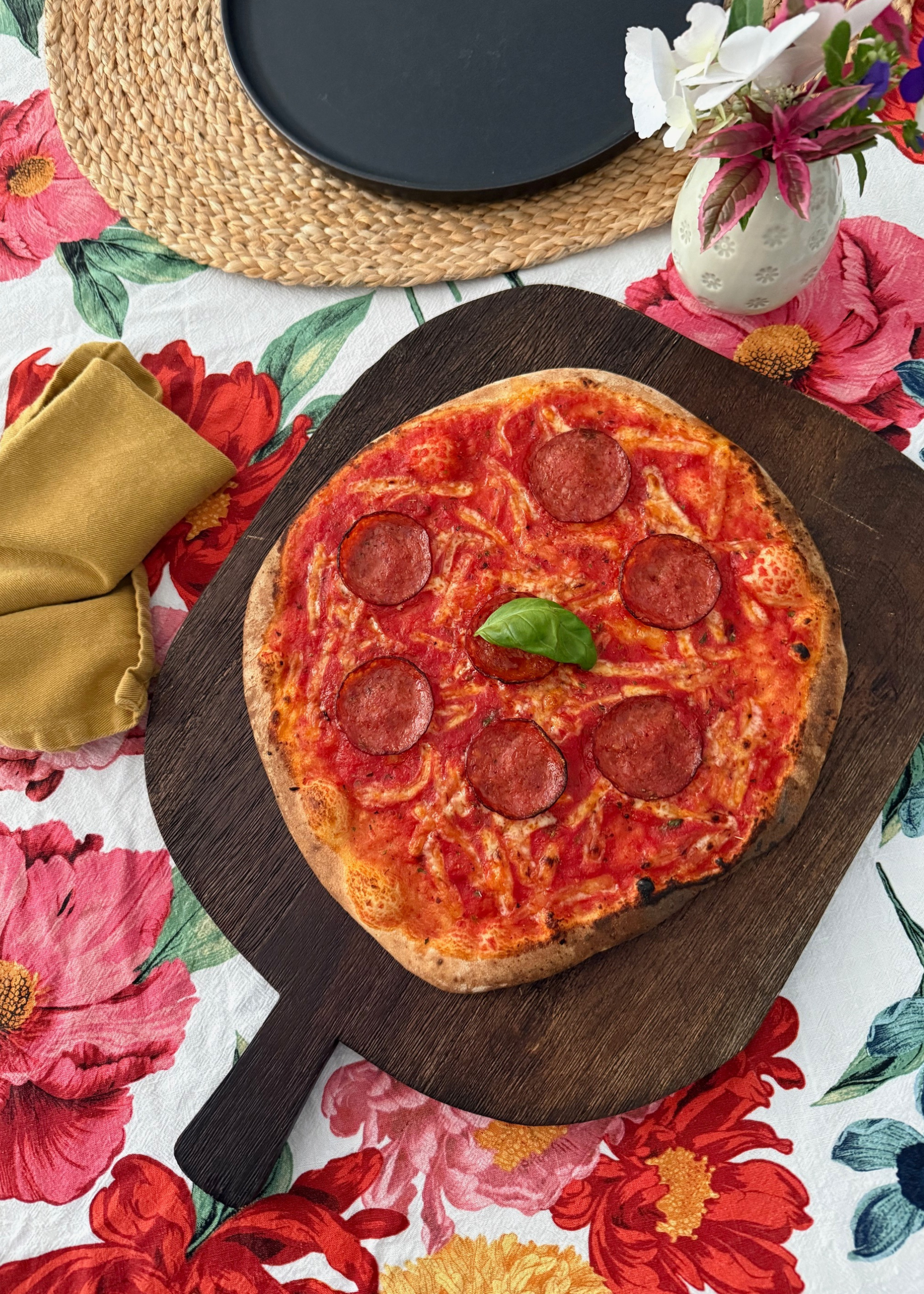 a whole pizza on a dark wood chopping board on top of a floral tablecloth next to a placemat with a black empty plate and a yellow napkin and a small vase of flowers