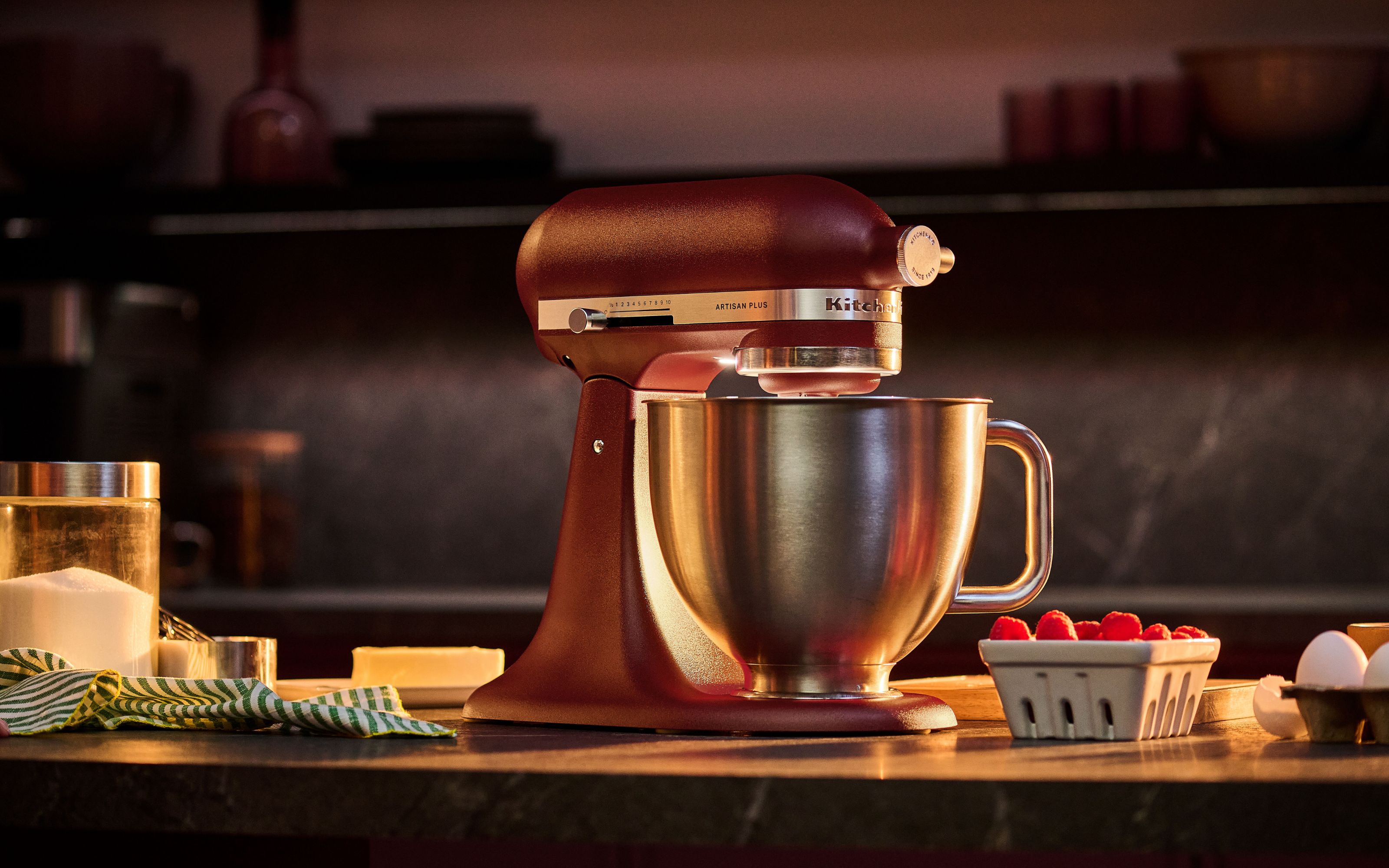 Red kitchenaid stand mixer on kitchen worktops with tea towel, bowl of sugar, raspberries and butter