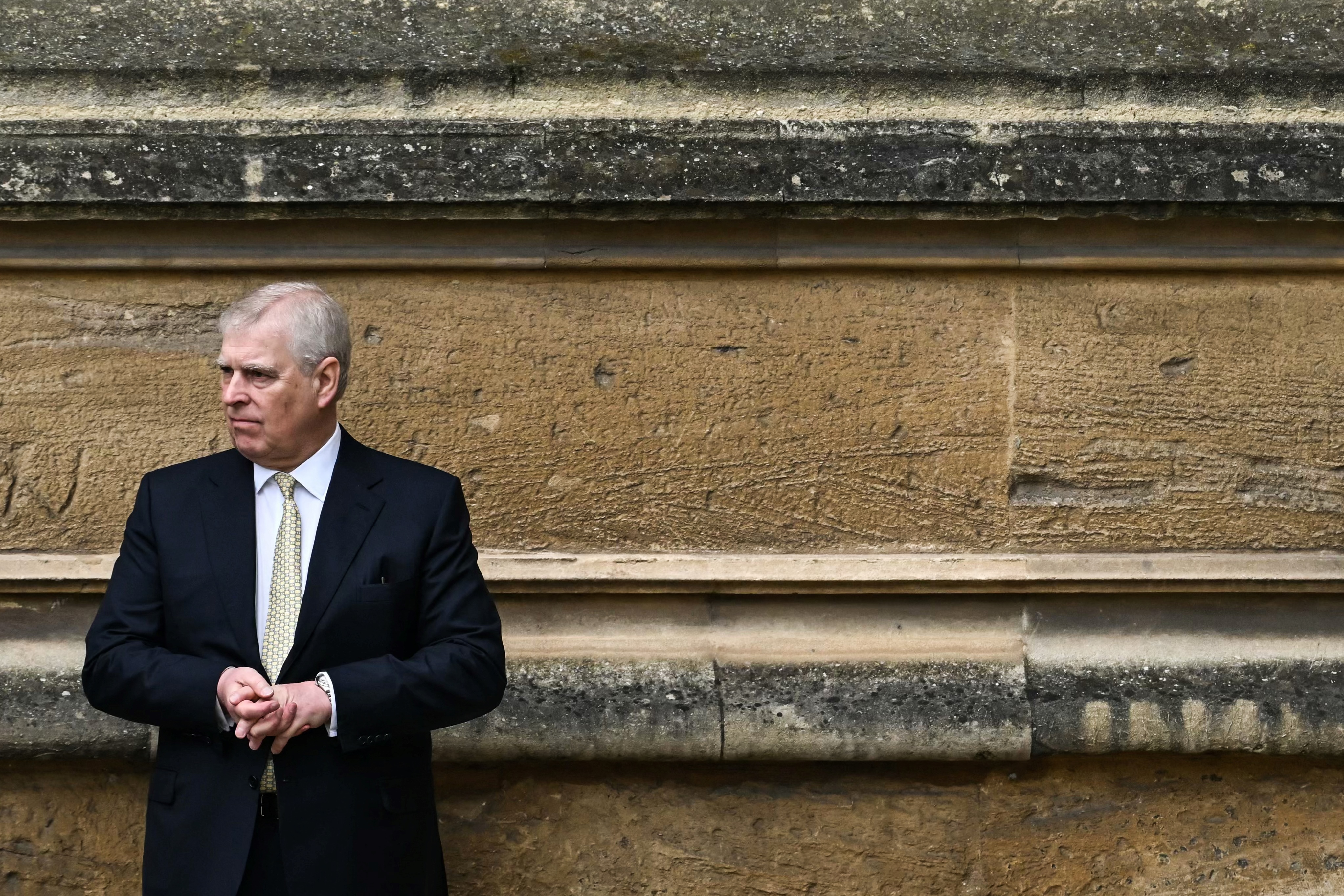 Prince Andrew standing against a stone wall in a suit and tie