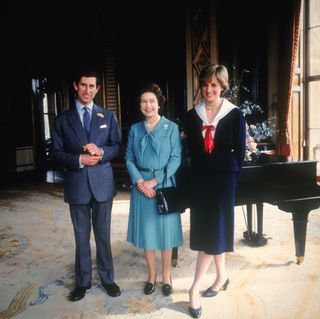 Prince Charles in a blue suit and Queen Elizabeth and Princess Diana wearing blue dresses standing in front of a piano on a patterned yellow and blue carpet