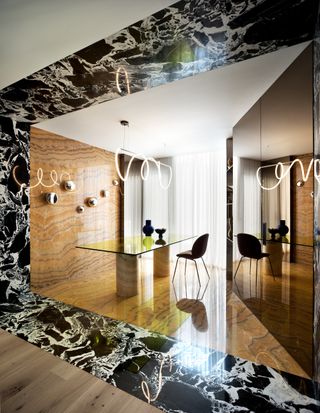 Image of an office space where the walls, floor, and legs of the desk are drenched in the same tan striped marble. The doorway of the room is a bold, black-and-white marble that connects to the tan marble in the room. There are silver objects hanging on the wall, and a neon light piece over the desk.