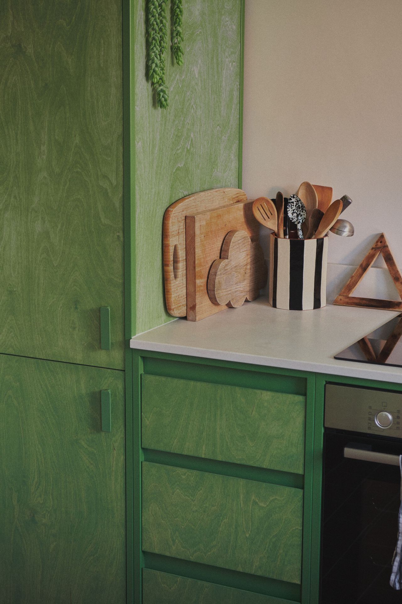 Image of a kitchen with green, wood-stained cabinetry. There are multiple cutting boards stacked on the white countertops and there is a black-and-white striped jar holding cooking utensils next to the stove.