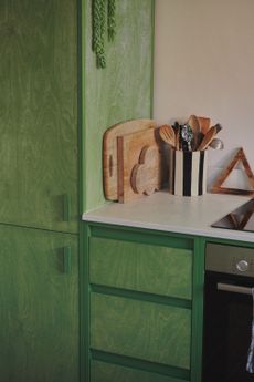 Image of a kitchen with green, wood-stained cabinetry. There are multiple cutting boards stacked on the white countertops and there is a black-and-white striped jar holding cooking utensils next to the stove. 