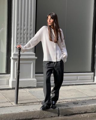 A woman wears a white button up shirt with gray trousers and black loafers on the sidewalk.