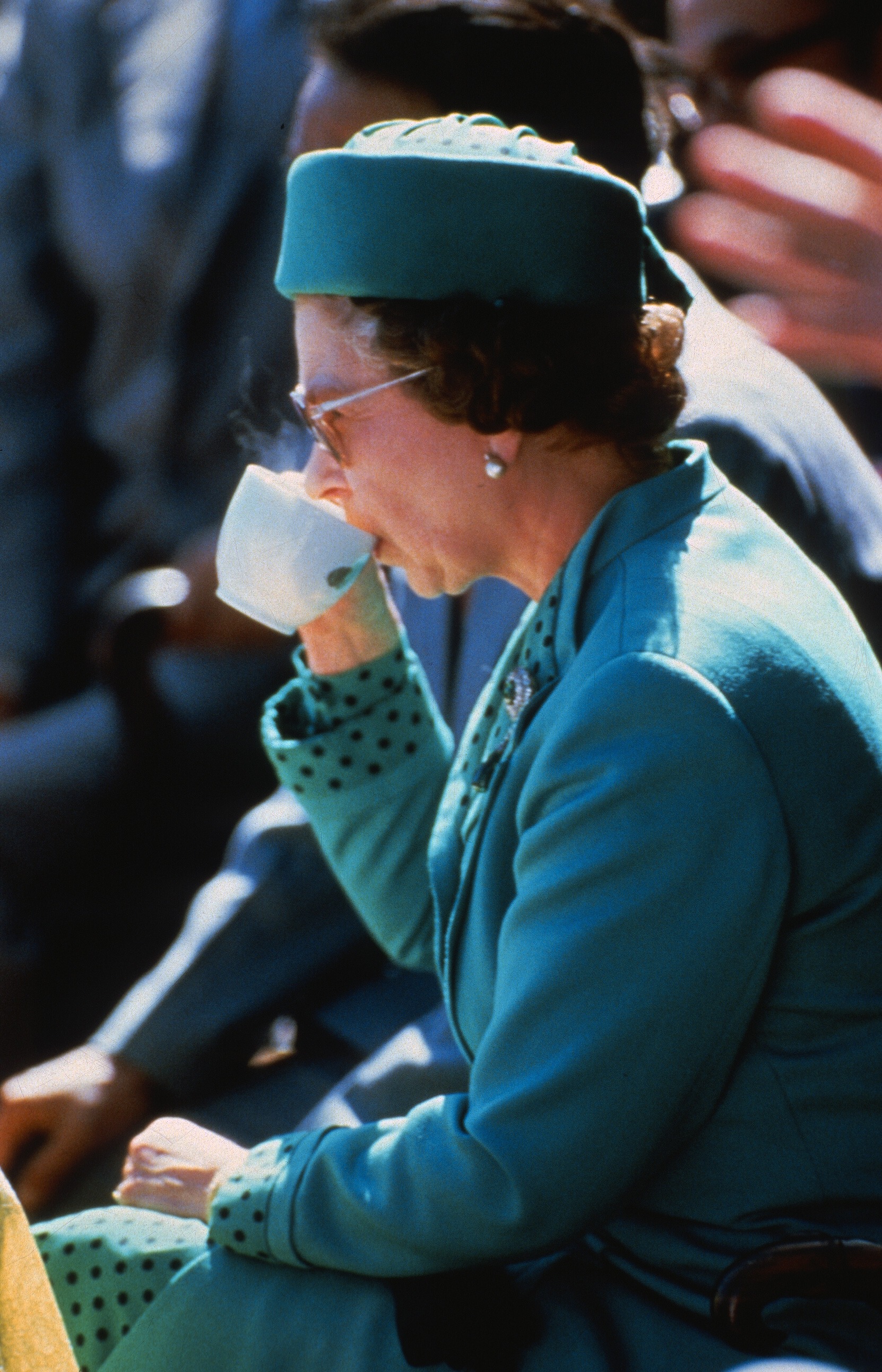 Queen Elizabeth drinking tea wearing a blue hat and coat
