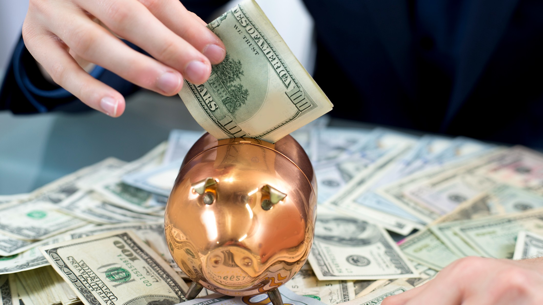 a hand slides a one hundred dollar bill into a gold piggy bank sitting on top of scattered hundred dollar bills