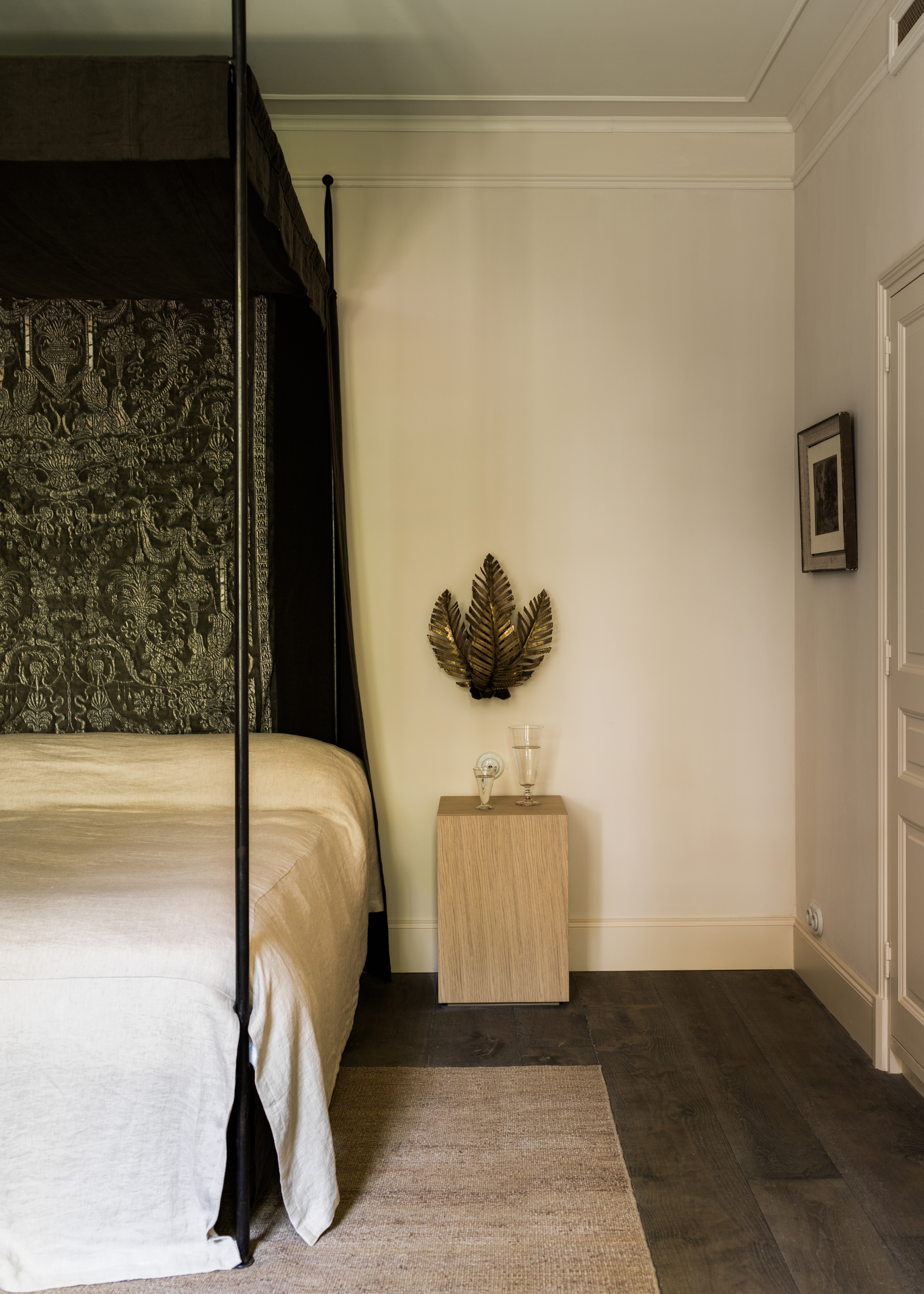 A white bedroom with a bed canopy, a white bed, a rug, a wood side table, and a textured wall sconce