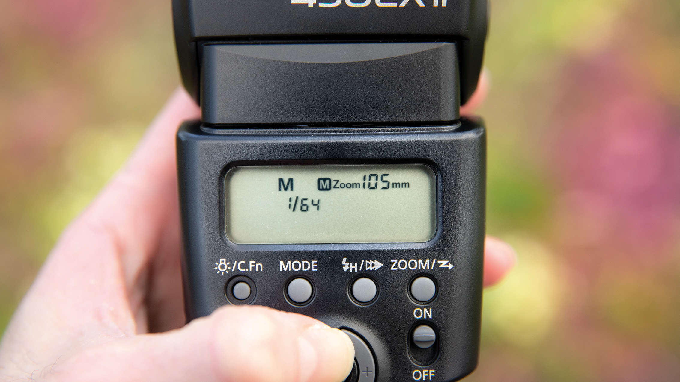 Close-up of a hand adjusting settings on a camera flash unit. The screen displays "M Zoom 105mm 1/64" with buttons for mode, zoom, and power. Blurred floral background