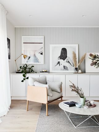 A light grey living room with marble coffee table, grey and wood armchair, wall art leant on white cabinets and gold table lamp