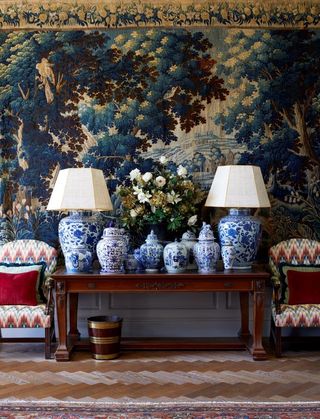 A hallway with a side table decorated with two lamps and blue china vases, and a large tapestry hanging on the wall