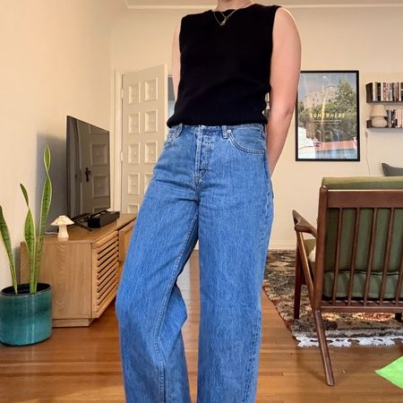 Woman wearing Alex Mill cashmere vest and Still Here jeans inside a mid-century modern living room. 