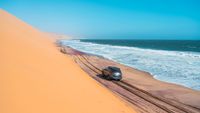 A silver sedan drives on a dark sandy road with a towering desert dune on the left and a blue ocean on the right