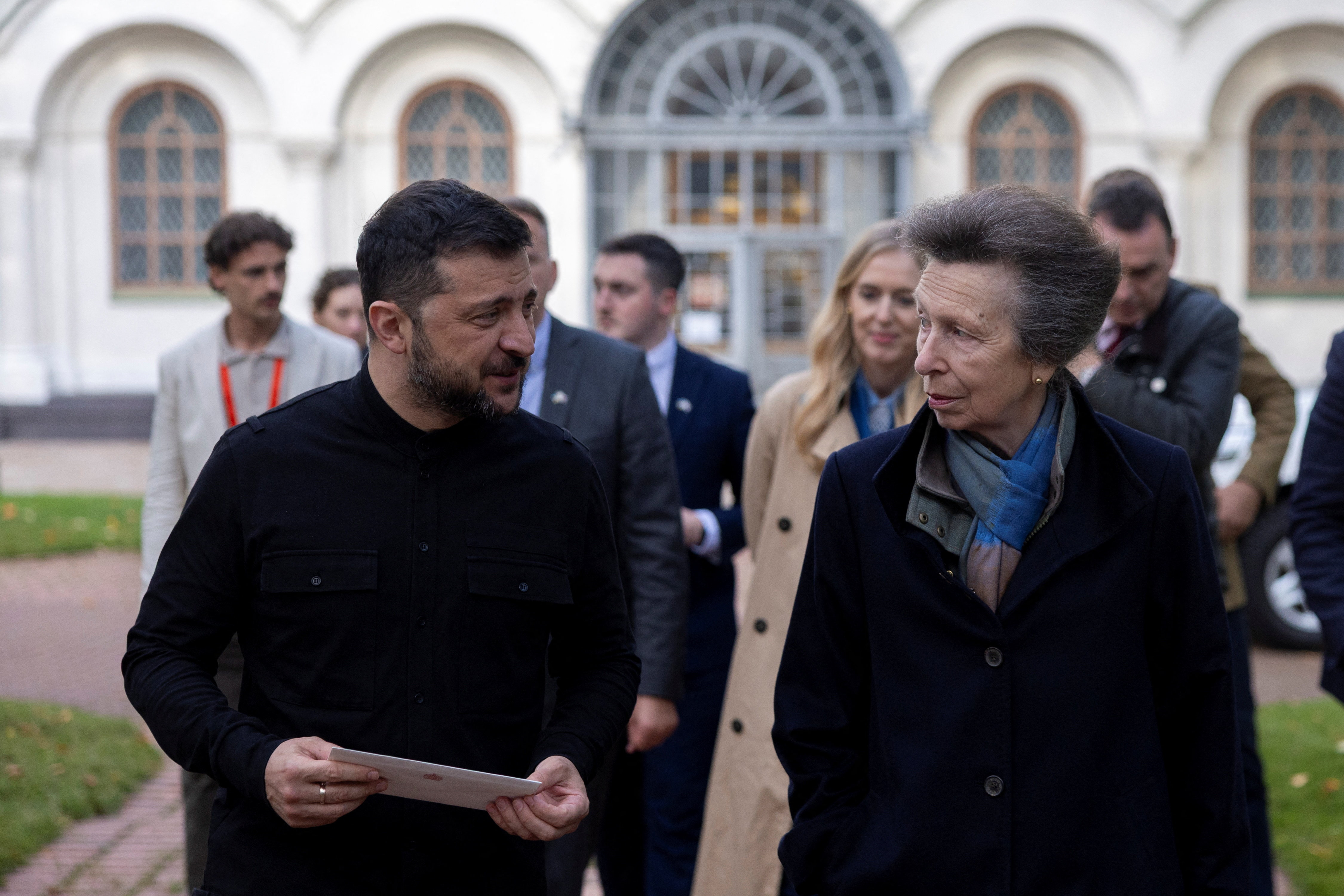 Princess Anne and Volodymyr Zelenskyy walking in front of a group of people outside a building with arched windows