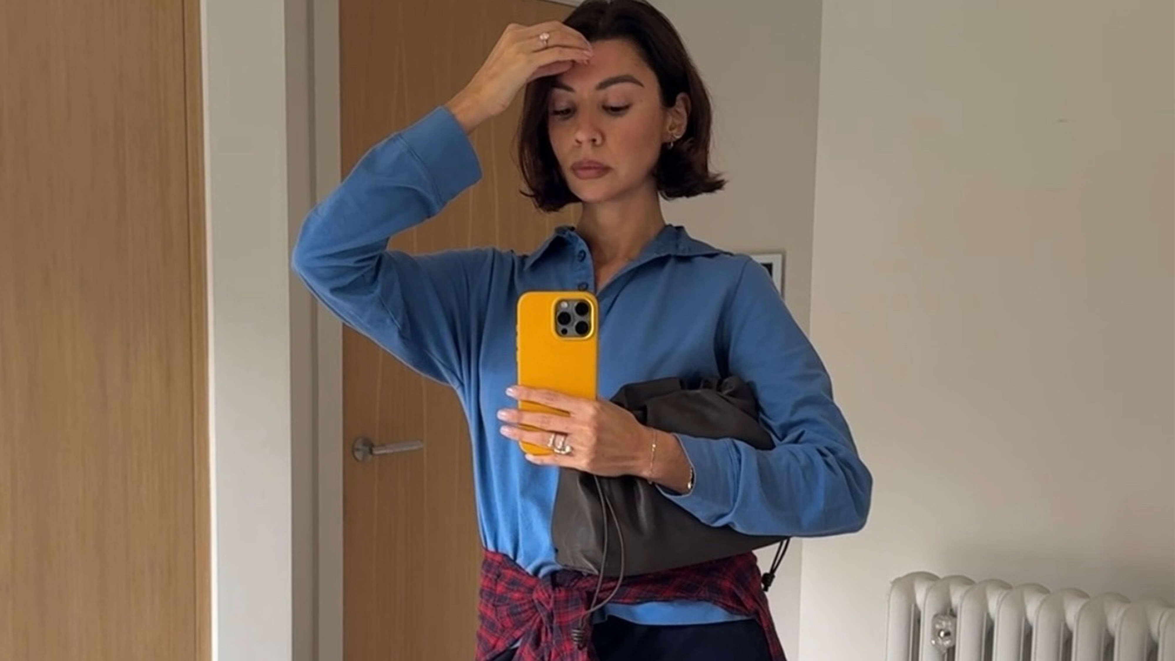 A woman wears a blue polo sweater, red plaid shirt, and holds a gray clutch.