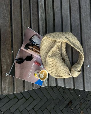 yellow braided bag next to coffee, magazine and lip gloss.