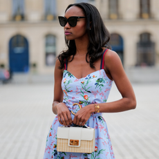 milie Joseph wears black large sunglasses, crystal earrings, a pale blue with red / green / navy blue / beige flower print pattern heart neck / tank-top / belted / midi dress, a beige wickers and white shiny leather bi-material handbag, during a street style fashion photo session, on April 29, 2023 in Paris, France. 