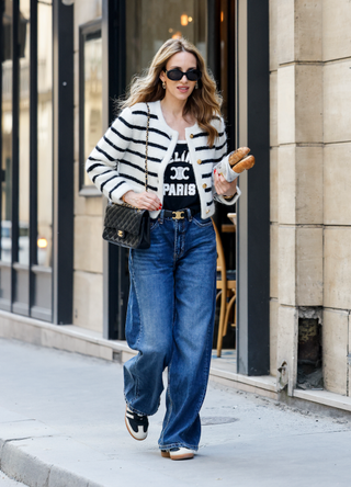 Alexandra Lapp, waering a blue top by Celine, a white jacket with white stripes by Celine, a black belt by Celine, a Jeans by Citizen of Humanity, blue Sneakers by Adidas, a black classic flap bag by Chanel and sunglasses by Celine, during a streetstyle shooting on May 12, 2024 in Paris, France