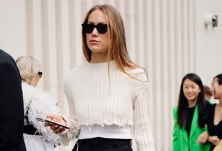 A street style photo from London Fashion Week showing a guest wearing an elegant cream cropped sweater with black Bermuda shorts and strappy heeled sandals.