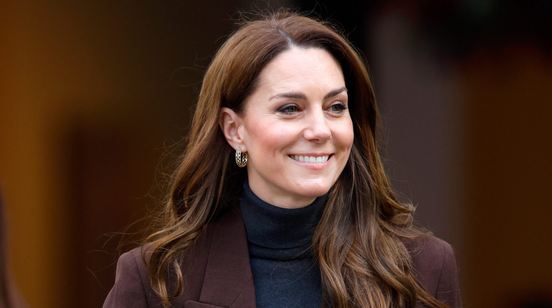 Princess Kate Middleton has long wavy brown hair and wears a black polo neck sweater with a brown blazer