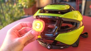 Lumos Ultra Fly Pro bike helmet in neon yellow against a red background. A user's hand is removing the magnetic taillight from the back of the helmet