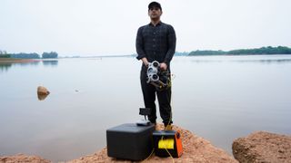 Chasing M2S Underwater Drone or ROV Underwater drone operator with the Chasing M2S with travel case, controller resting on the case, and the tether reel – plus the operator stood on a reddish rock by a lake.