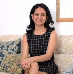 Aditi Sharma Maheshwari is a woman with dark brown, shoulder-length hair and a medium-olive skin tone, wearing a high-neck tweed dress and sitting in a neutral living room on a subtly patterned sofa