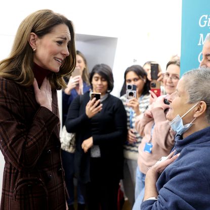 Kate Middleton wearing a plaid coat with her hand on her chest looking emotional talking to a woman with a mask pulled down and people. in the background taking photos at a hospital