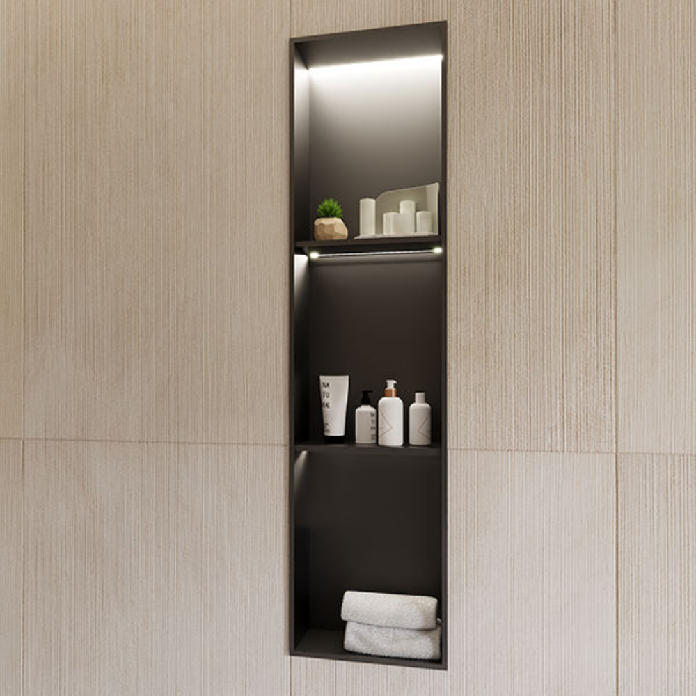 A tall, narrow recessed wall niche with a matte black interior and integrated LED strip lighting, set into a textured cream wall. Three open shelves display toiletry bottles and a small plant on the upper levels, with a neatly folded white towel on the bottom shelf.