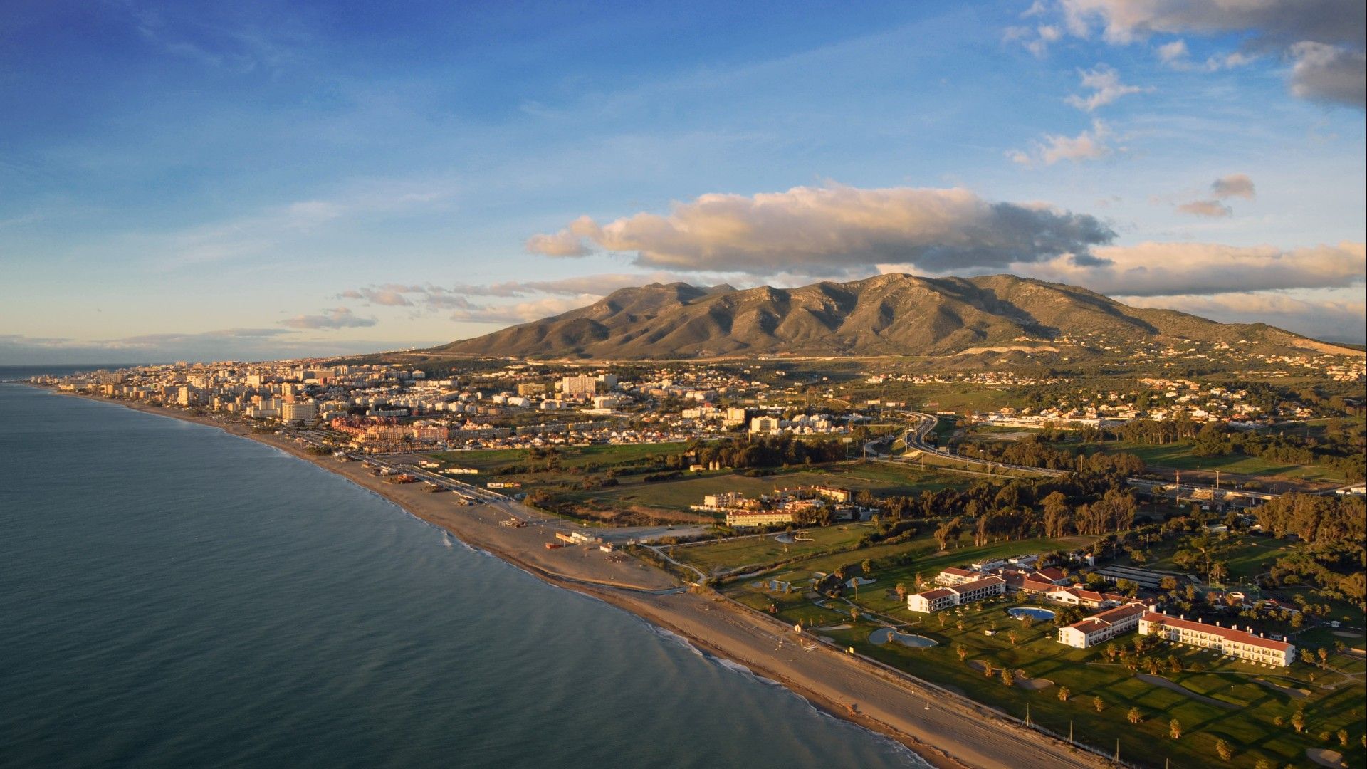 Aerial view of the Mediterranean coast. There are large hills in the background, looming over the city below. Above is a blue sky, dotted with large clouds.
