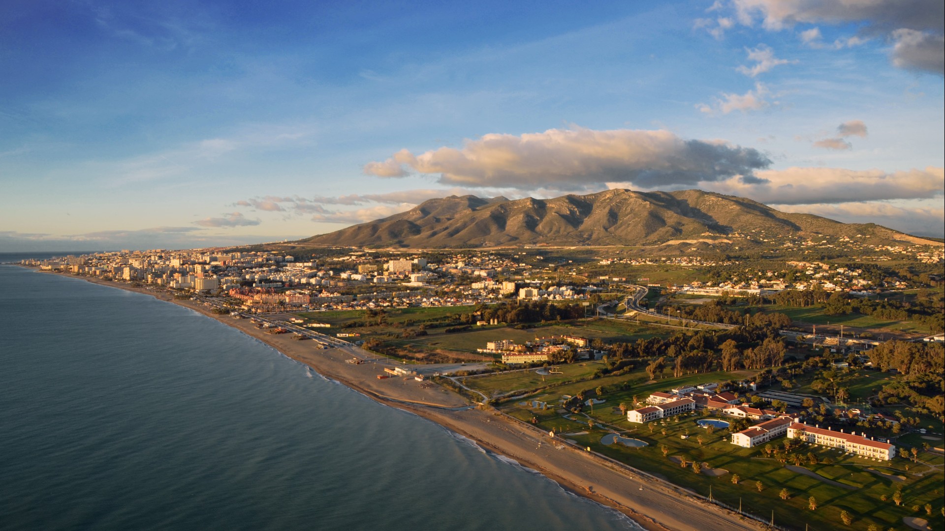 Vista aérea da costa mediterrânica. Há grandes colinas ao fundo, pairando sobre a cidade abaixo. Acima está um céu azul, pontilhado por grandes nuvens.