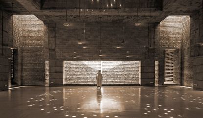 Interior view of a prayer hall at Bait ur Rouf Mosque, Bangladesh designed by Marina Tabassum. The prayer hall features exposed brick walls, tile flooring and multiple pendant lights. There is a man dressed in white standing in front of an illuminated wall that has another wall in front with a rectangle cut out design