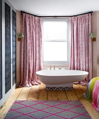 Freestanding bath with raised tiled stand in the bay window of the main bedroom, pink curtains, two wall lights with green shades, wooden flooring with pink patterned rug, multi-colored bedding