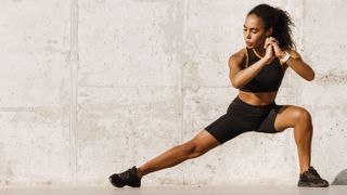 Woman in black workout clothes performing a side lunge on her left leg outdoors against a concrete background
