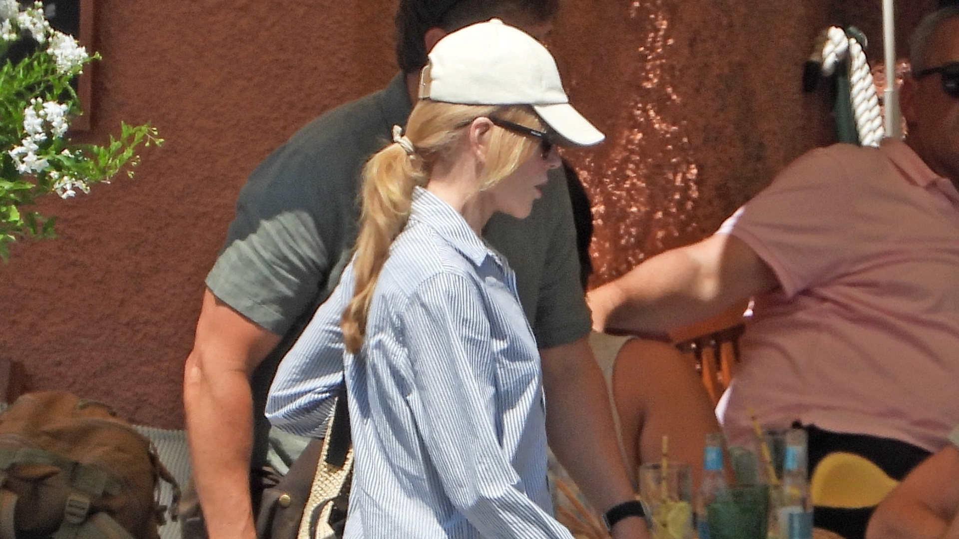 Nicole Kidman in Portofino wearing a white baseball cap, blue-and-white-striped button-down shirt, white cut-off denim shorts, a Loewe raffia shoulder bag, and white leather Adidas Samba sneakers