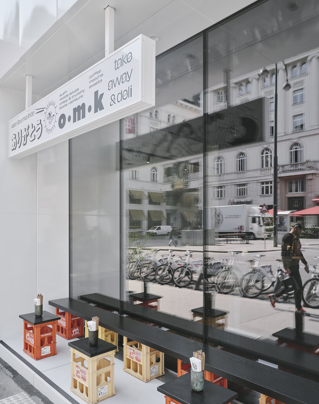The street exterior of a Japanese cafe features industrial-style benches and tables in wood and yellow or orange plastic, a white, hanging solid sign, and glass windows.
