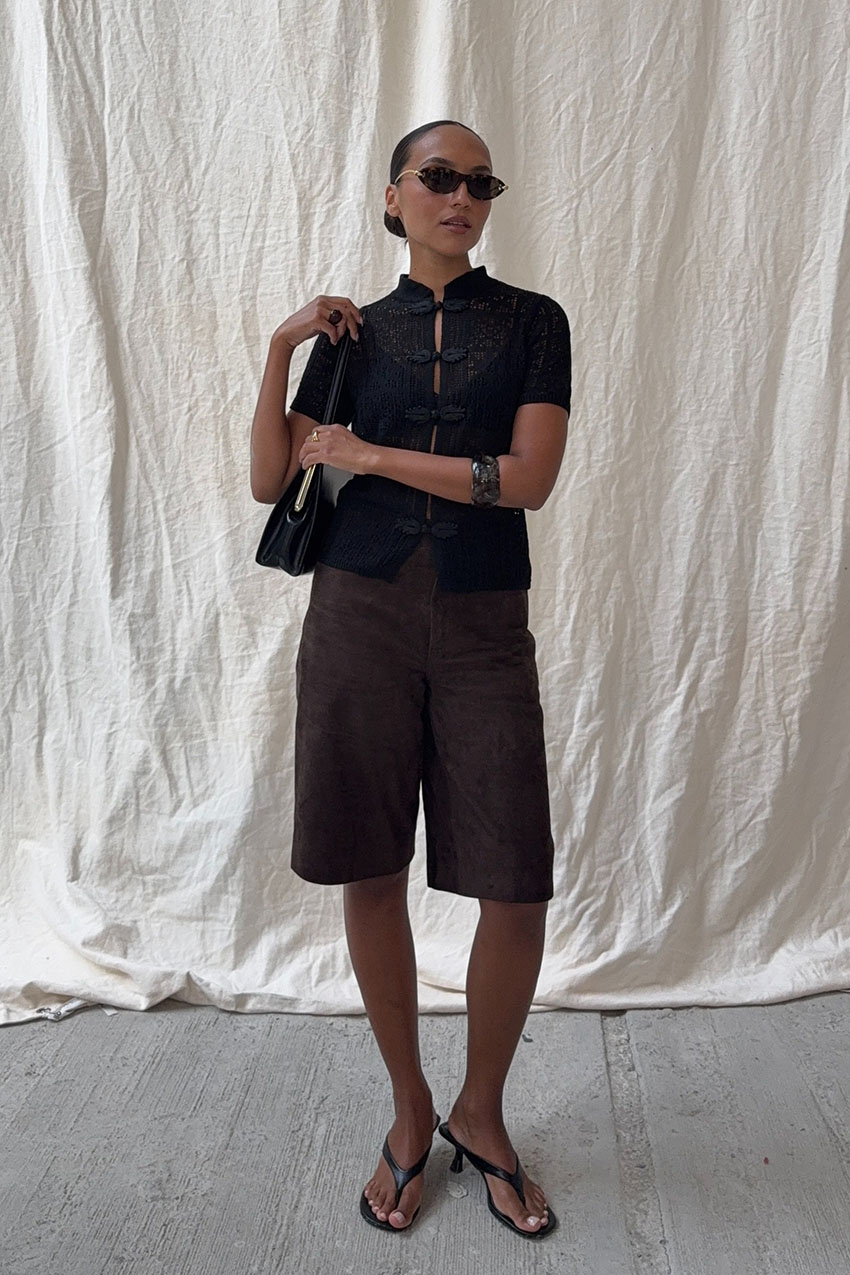 A woman wearing a black top, long brown shorts, black kitten heels, a black bag, and black sunglasses.