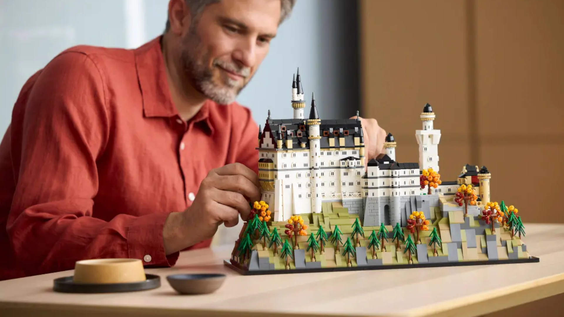 A man in a red shirt smiles and holds the Neuschwantstein Castle Lego set, which is standing on a wooden table