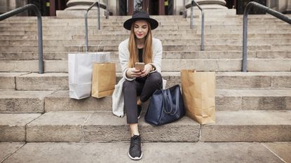 A young woman sits on the steps of a building, surrounded by shopping bags, while she looks at her phone.