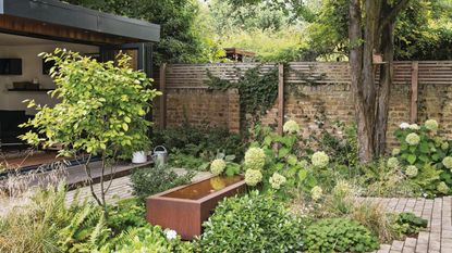 Garden with garden room, water feature patio paths and flower beds of shrubs and grasses. 