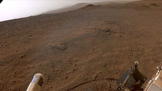 parts of a mars rover are seen in the foreground, with a desert landscape and small peaks visible in the background