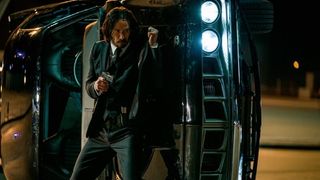 Keanu Reeves takes cover behind a flipped car in John Wick: Chapter 4