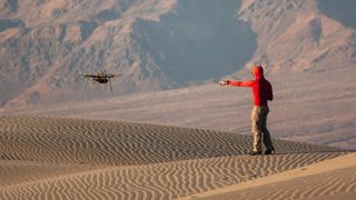 A man wearing a red hoodie and tan pants points to a nearby drone hovering over rippling sand dunes