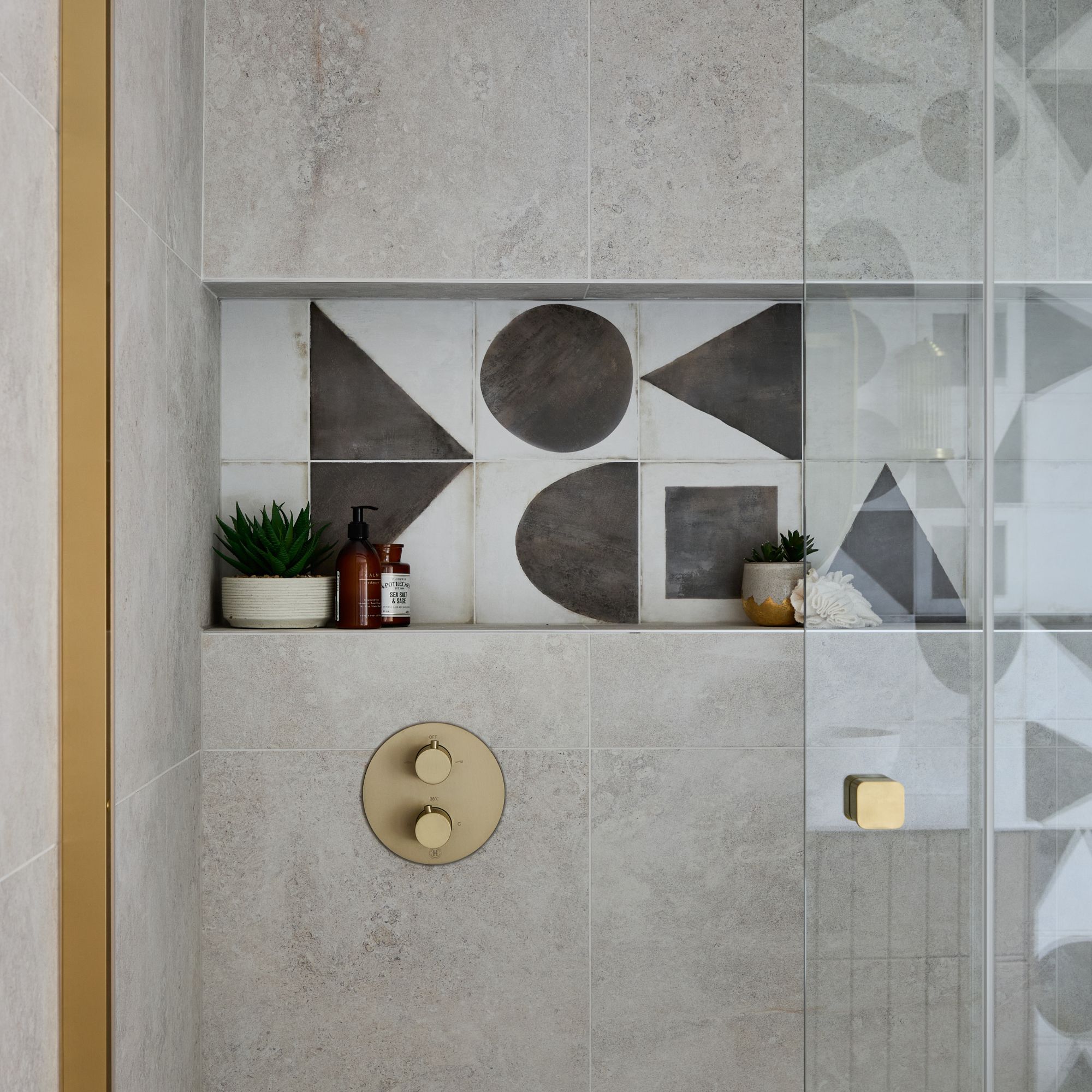 Shower area with large grey tiles, gold fittings and handles, recessed shelving with monochrome tiles