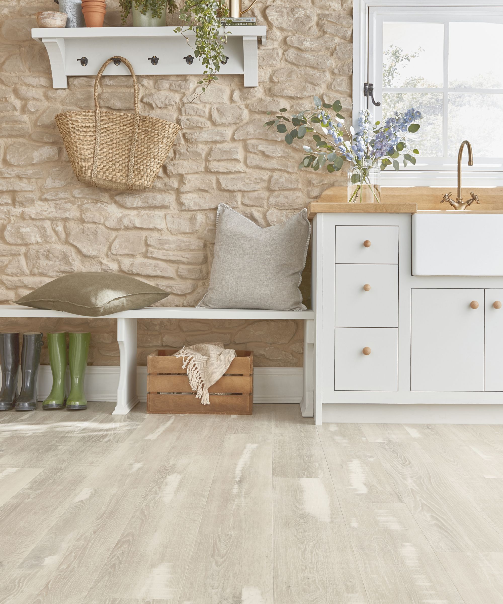 Charming country-style entryway with light wood-effect flooring, a stone wall, white cabinetry, and a white bench with pillows. Decorative touches include plants, rain boots, and a woven basket.