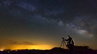 A man sits behind a camera mounted on a tripod with the night sky in the background