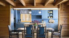 A dining room with wooden panelled walls and a view into a kitchen with blue zellige tiles and blue cabinetry