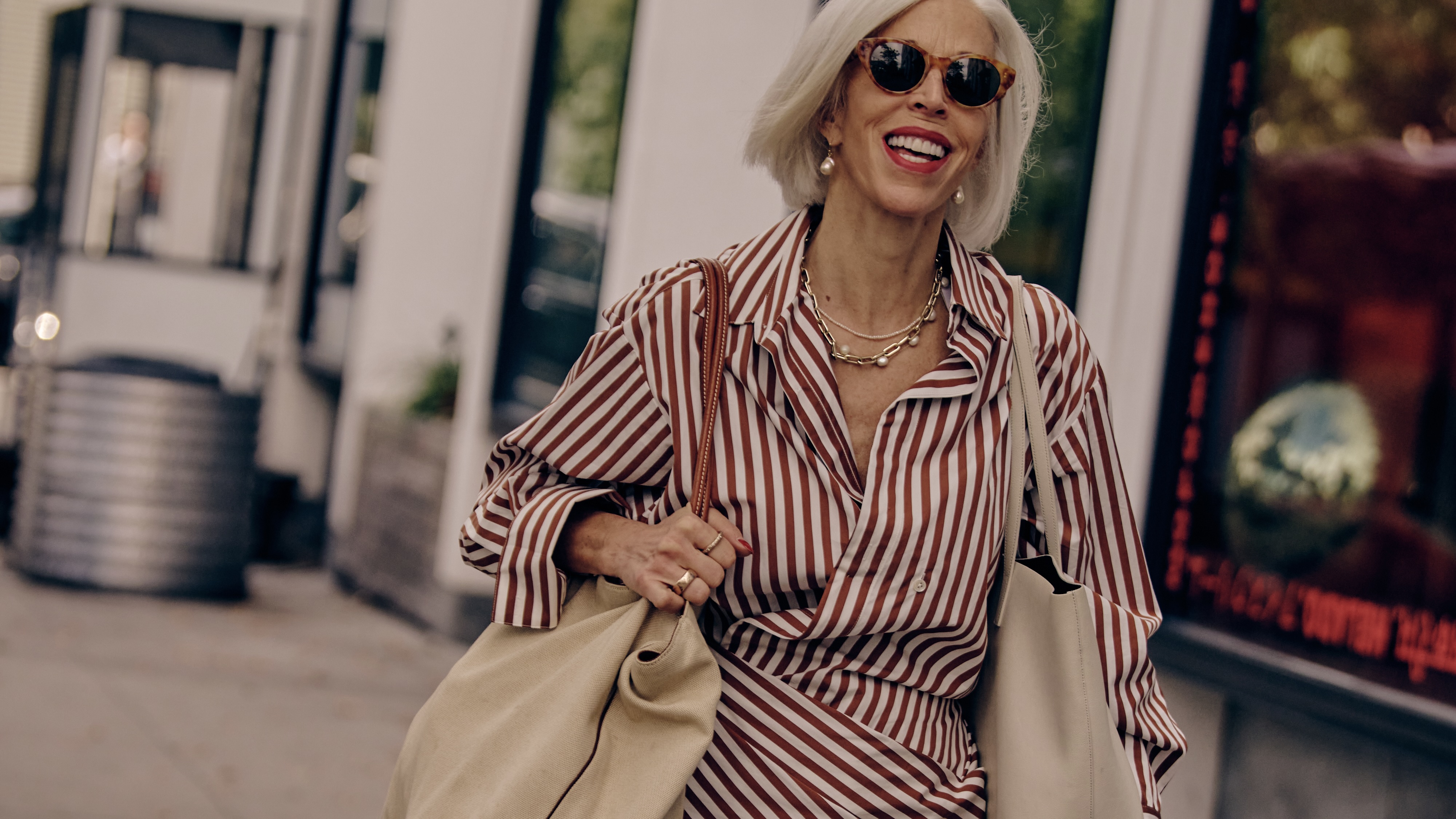 woman wearing a red and white striped poplin dress carrying a tan tote bag with sunglasses on