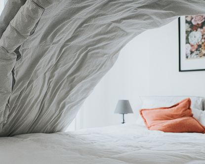 An action shot of someone making the bed in white bedroom with grey duvet, grey lampshade lighting decor, orange upholstered cushion and framed floral wall art