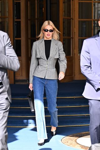 Kendall wears black rectangular sunglasses, a black t-shirt, gray cinched blazer, blue straight-leg jeans, a black bag, and black ballet flats.